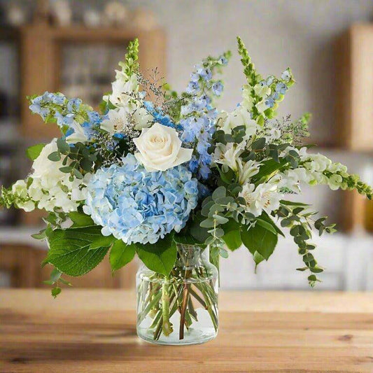 Blue and white floral arrangement with white and blue roses, stock and hydrangeas in a clear vase.