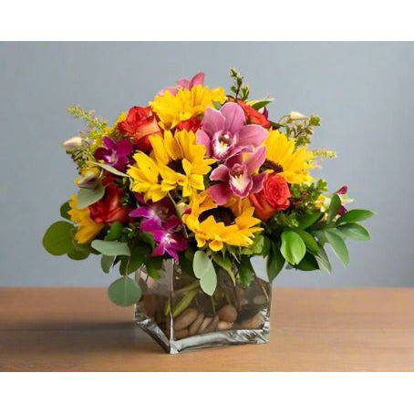A floral arrangement with sunflowers roses and fillers with greens in a square clear vase.