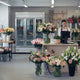 A flower shop with a modern design. with various floral arrangements displayed in vases. There's a stand with a woman employee using the cash register. In the background, there's a cooler with three doors. The shop has a light and airy atmosphere