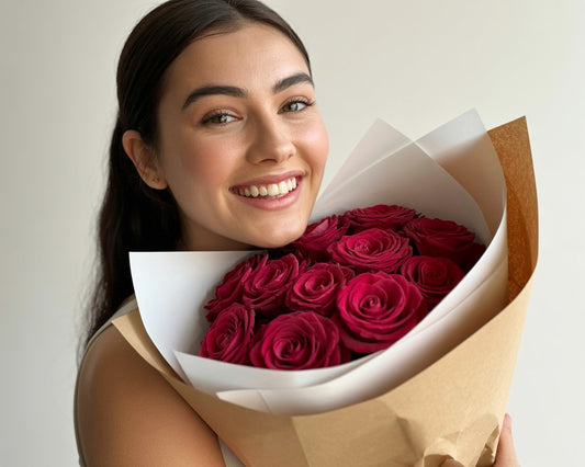A young woman facing the camera with a warm, genuine smile radiating pure happiness. She holds a carefully wrapped bouquet of deep crimson red roses against her chest.