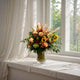 A vibrant flower arrangement in a clear glass vase featuring sunflowers, peach and red roses, and lush greenery, placed on a windowsill with soft natural light coming through white curtains.