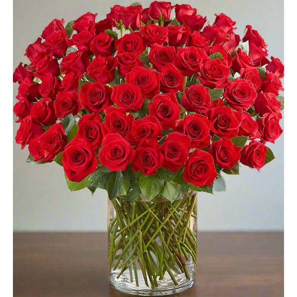 100 Fresh Premium Long Stem Red Roses in a clear glass vase set in a wooden table. 