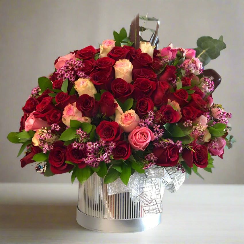 A bouquet of red, pink, and white roses with green leaves arranged in a silver container.