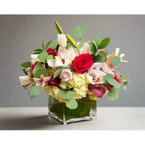 Floral arrangement with pink and red roses eucalyptus and fillers in a clear square vase.