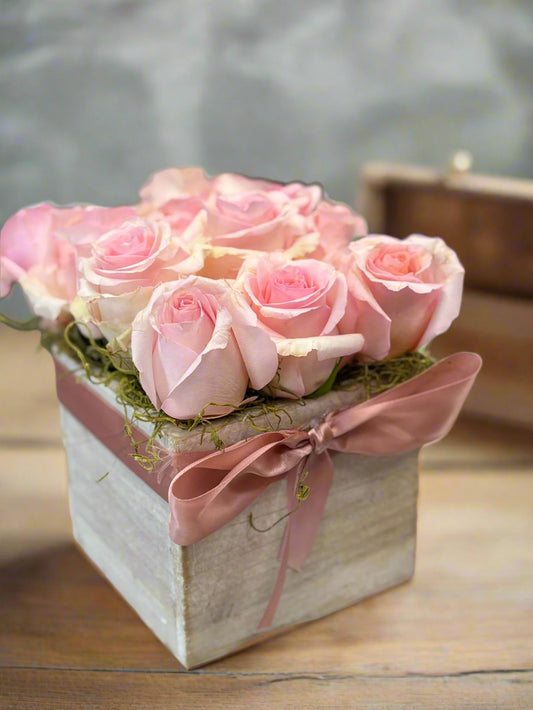 Light pink roses in a wooden box with ribbon, Mother’s Day flower arrangement on a table.