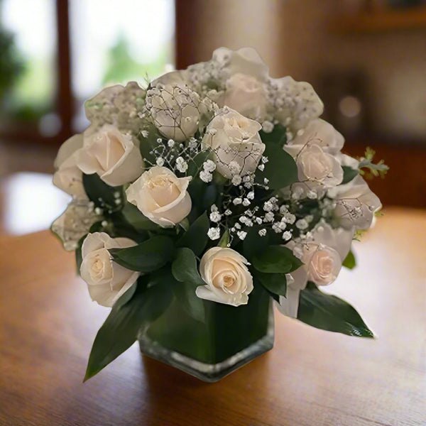 18 White Rose Arrangement in a clear square vase, et in a wooden table.