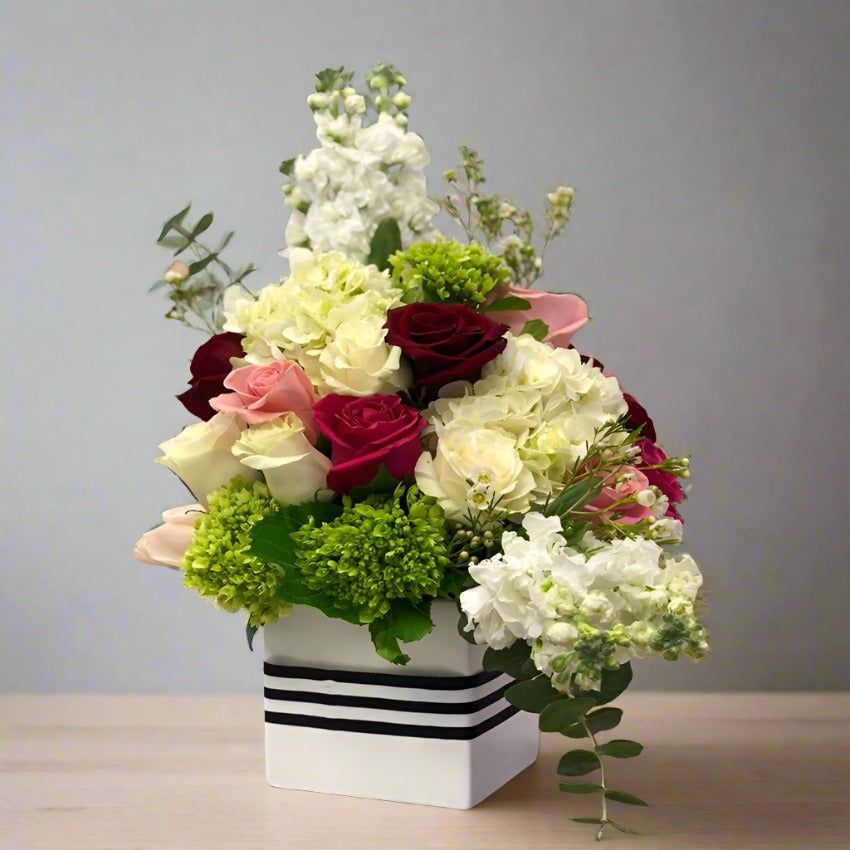 A bouquet of mixed flowers including red and white roses in a white vase with a black stripe.