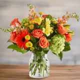 fall floral arrangement in a clear vase with orange, yellow, red and green flowers.