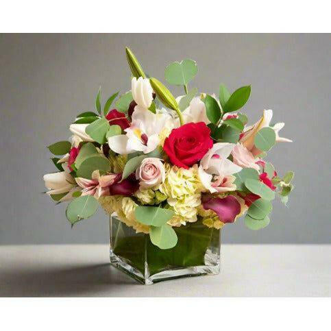 Mixed floral arrangement with red yellow and white flowers with lost of greens in a square clear vase.