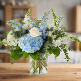 Bouquet of white roses, blue hydrangeas, and assorted flowers in a clear glass vase on a wooden table.