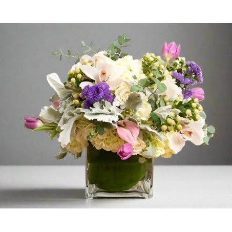 A floral arrangement in a clear glass vase featuring white, pink, and purple flowers with green foliage.