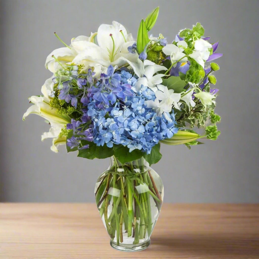 Bouquet of white lilies and blue hydrangeas in a clear glass vase on a wooden table