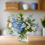 A vase of blue and white flowers arranged on a wooden table with a blurred background of shelves and containers.