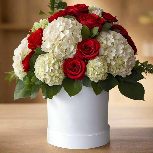 A bouquet of white hydrangeas and red roses in a white cylindrical vase on a wooden table.