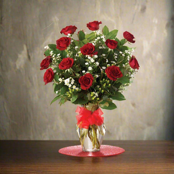 A bouquet of red roses arranged in a clear vase with a red ribbon, placed on a wooden table.