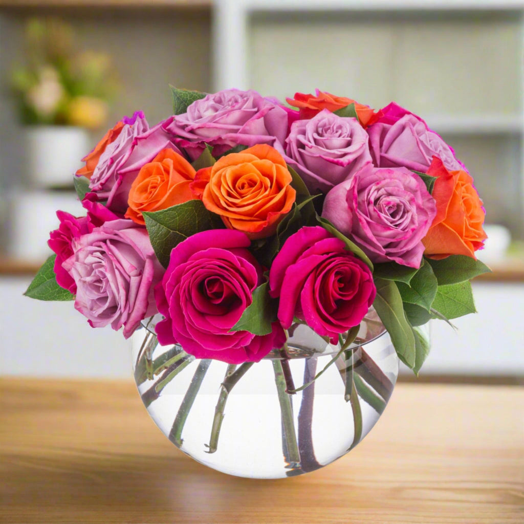 A round glass vase filled with bright pink, purple, and orange roses, placed on a wooden table.