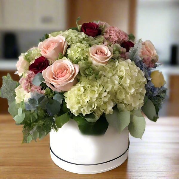 Bouquet of mixed flowers including pink and red roses, white hydrangeas, and green foliage in a white vase on a wooden table.