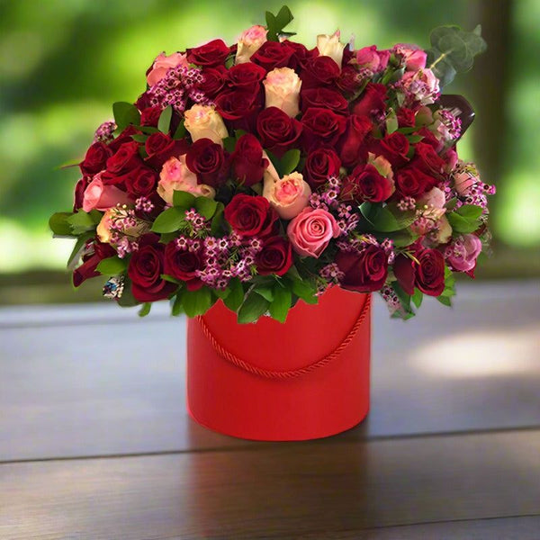 Bouquet of red, pink, and white roses in a red round box on a wooden table.