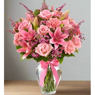 A bouquet of assorted pink flowers in a clear glass vase with a pink ribbon.