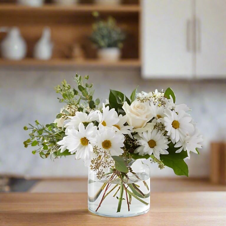 A bouquet of white flowers in a clear glass vase, placed on a wooden table in a kitchen setting.