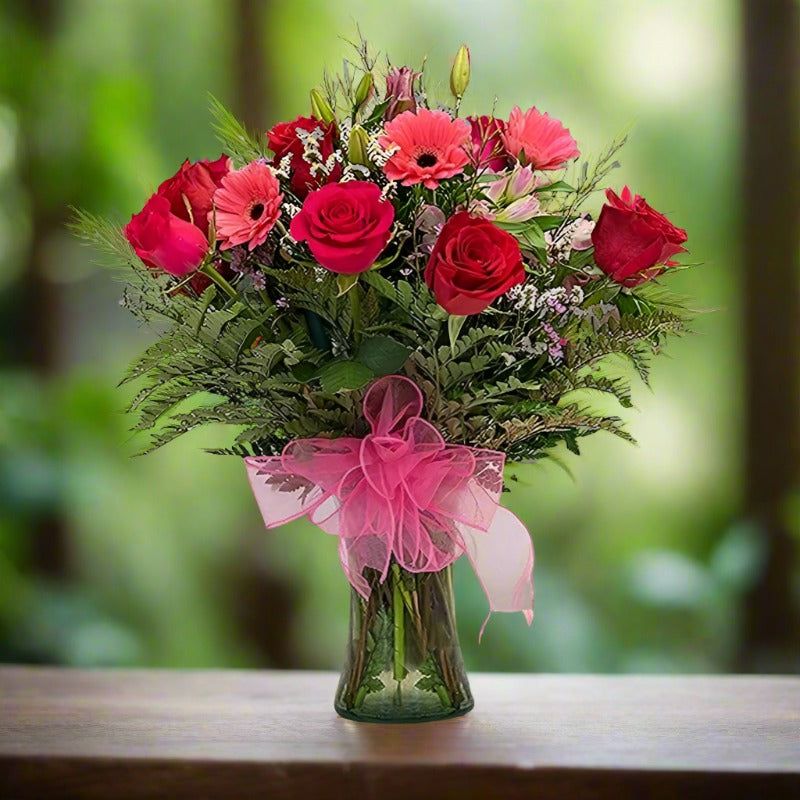 A bouquet of red and pink flowers in a green vase with a pink ribbon, set against a blurred green background.