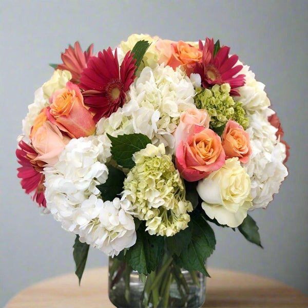 Bouquet of mixed flowers in a glass vase, including roses, hydrangeas, and gerbera daisies.