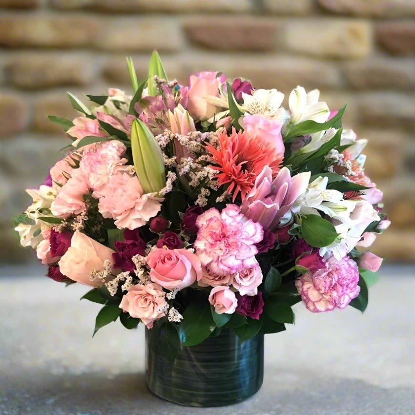 A bouquet of assorted pink and purple flowers in a green vase, set against a stone wall background.