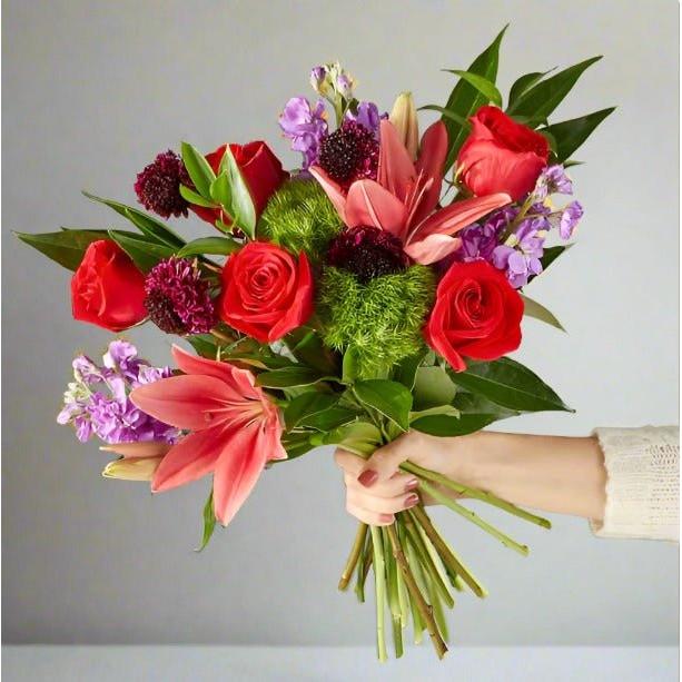 A hand holding a bouquet of colorful flowers including red roses, pink lilies, and purple blooms.