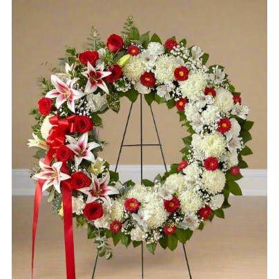 Red and White Standing Wreath with roses, lilies, pomp's and a red ribbon on the side.