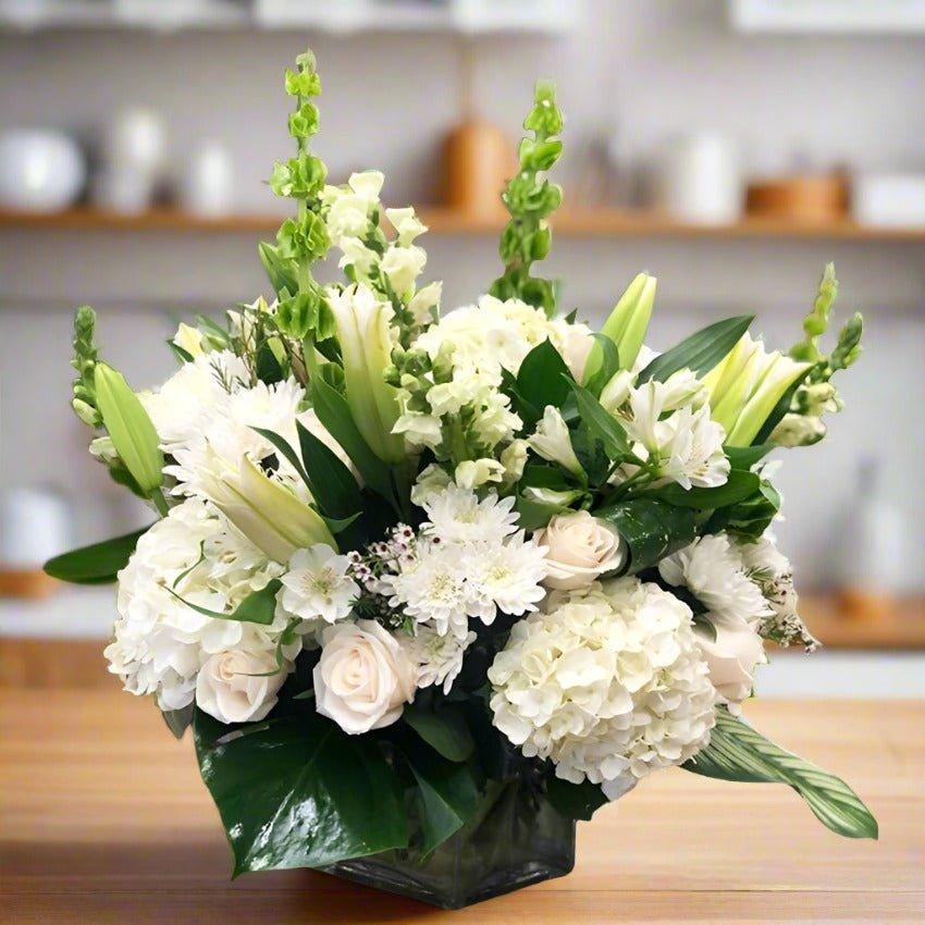 A fresh floral arrangement with white roses, lilies, and greenery in a glass vase on a wooden table.